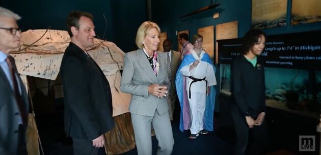 Betsy DeVos tours a school while Torin Hodgman wears a trans flag in the background.