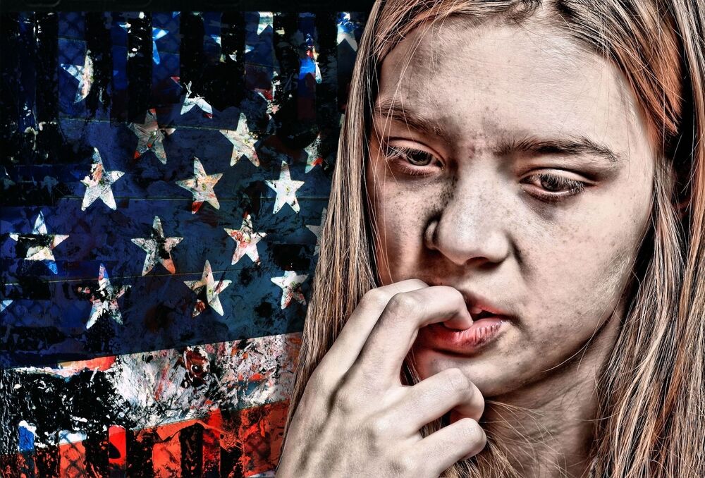 A young white woman bites her nails in front of a ragged and dark American flag