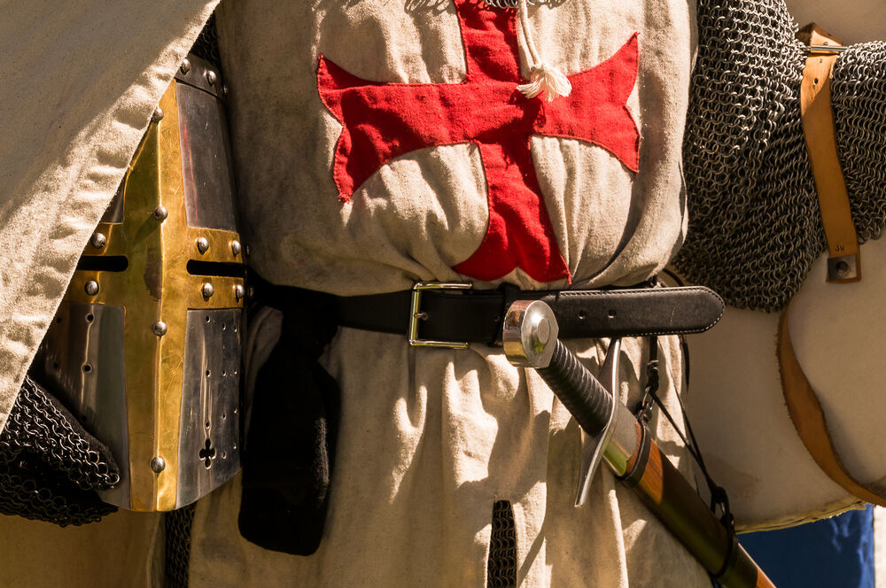 A close shot of persons torso dressed up historically to mimic a knights templar in full armour holding his helmet under the arm with his sword visible in the belt.