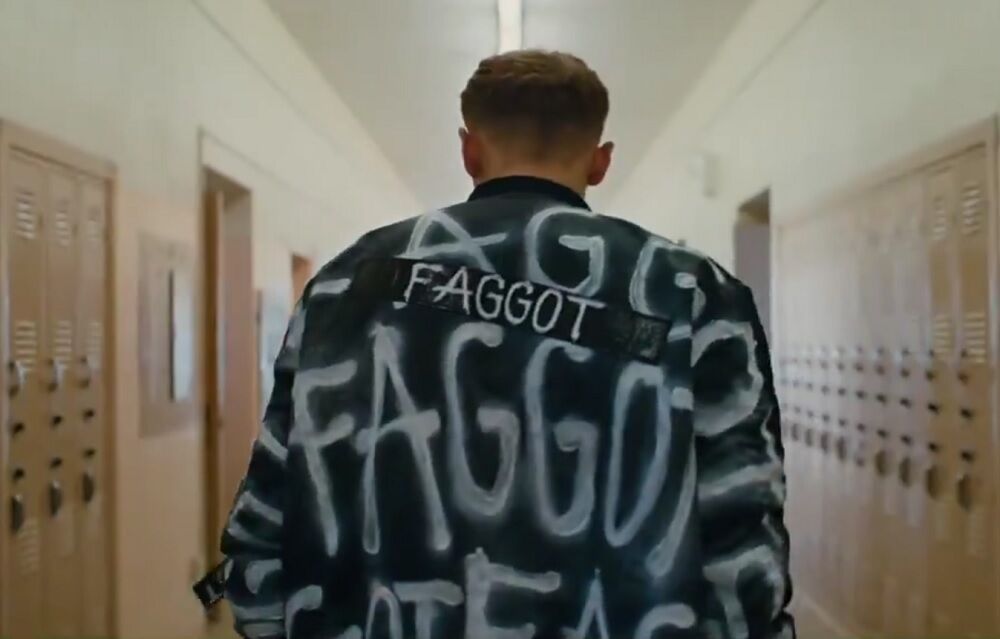 A white person wearing a black jacket with the word "faggot" written on it.