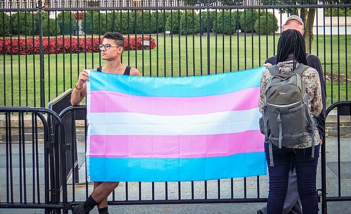 Two young people holding the blue, pink, and white transgender flag in front of the White House.