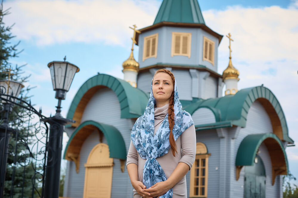 A Russian woman stands in front of a small wooden church.