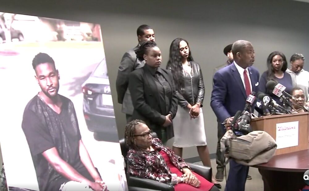 A press conference with a lawyer at a lectern and the family of Jonathan Hart behind him. There is a large image of Jonathan Hart.