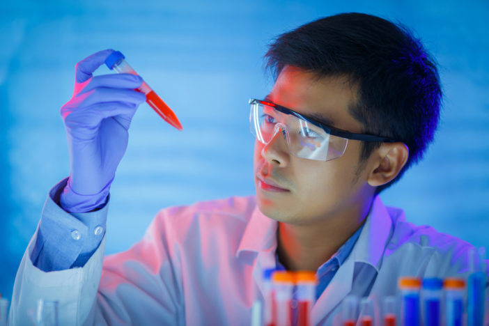 A man with gloves and lab goggles looks at a test tube with red liquid in it.