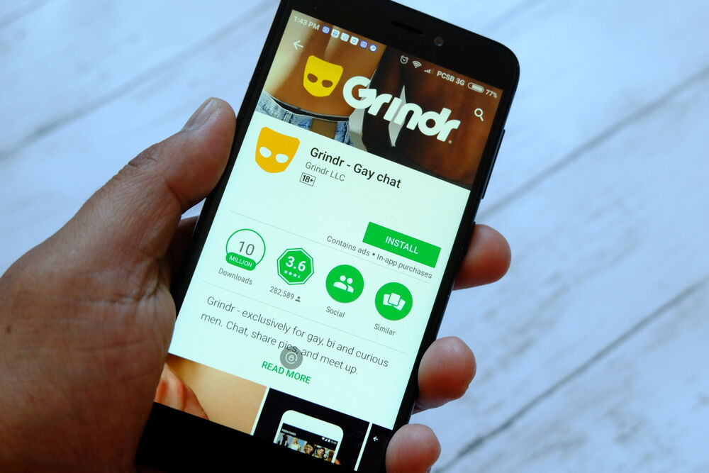 A black hand holds a smartphone showing a Grindr grid