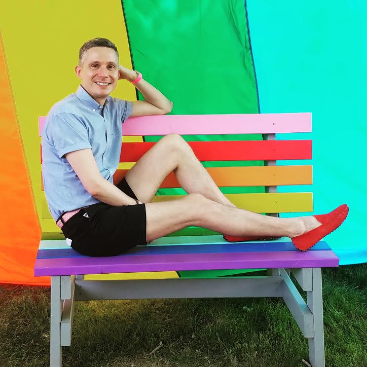 Gay Macy's employee Matt Ossenfort poses in a blue polo and shirts on a rainbow bench, against a rainbow backdrop.