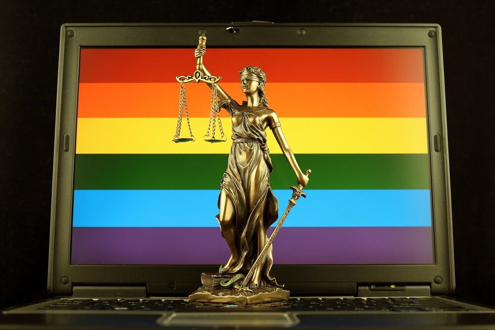 A computer with a rainbow on it and Justice, the statue, is in front of it. Perhaps it's a symbol of online, blind, queer justice, or maybe it's a satirical statement about social justice keyboard warriors. It's a stock photo, confusing in its symbolism. Also the laptop in the pic looks like it's from 1996.