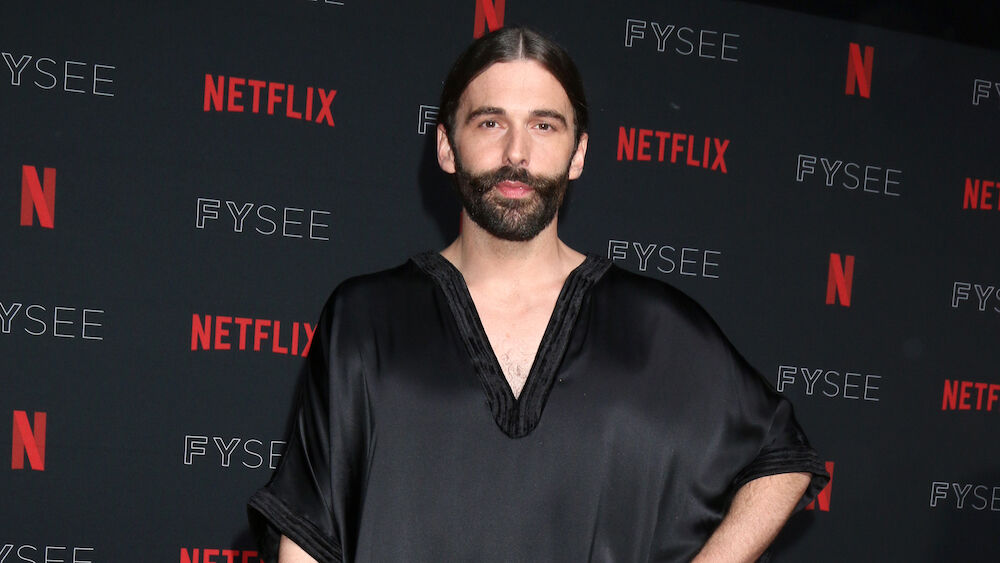 Jonathan Van Ness, a bearded white person with long hair, stands in a black shiny dress at a Netflix red carpet event.