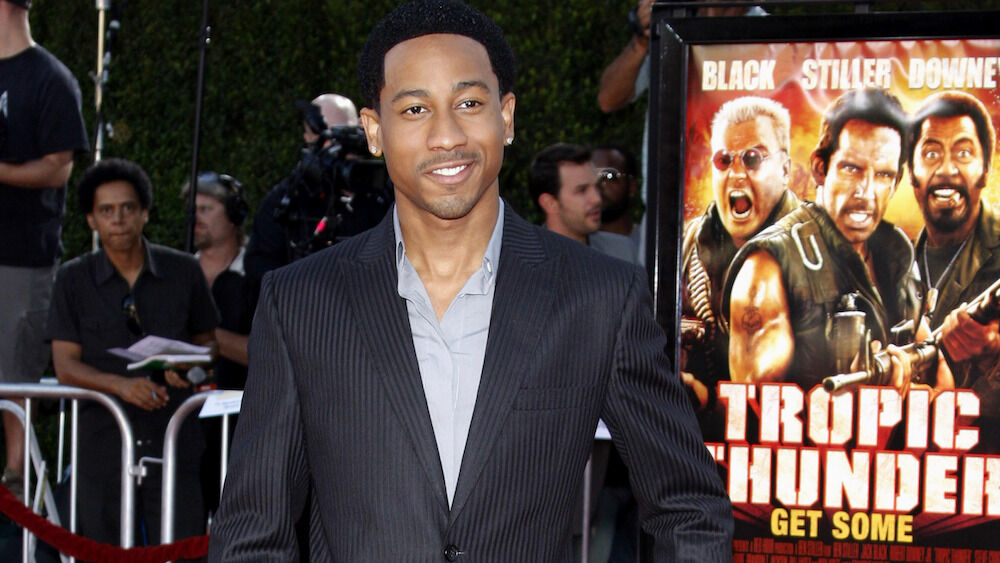 Brandon T. Jackson wears a suit and stands next to a "Tropic Thunder" movie poster