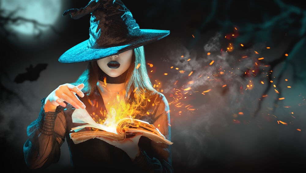Beautiful young woman in witches hat conjuring, making witchcraft by making flames erupt from the pages of a book of magic during a full moon..