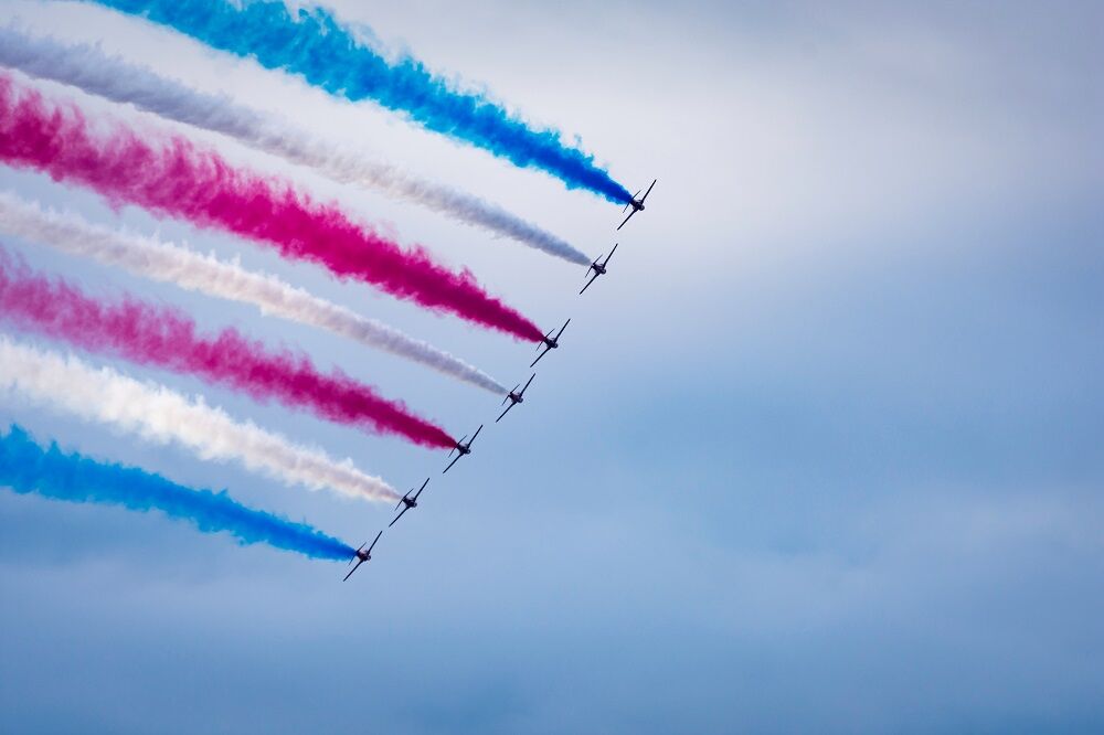 Aircraft performance and formation of 7 planes banking to form a stunning curved red blue and white smoke trail against a cloudy sky with side by side arangement