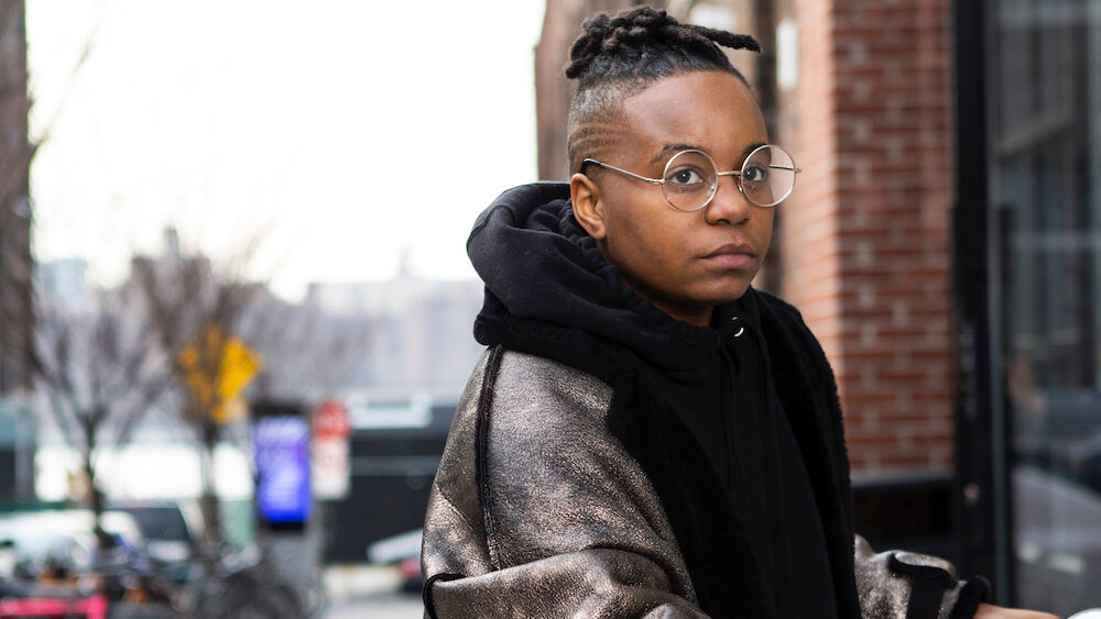 A black transgender man looks at the camera while parking his vehicle outside a brick building on a cold day.