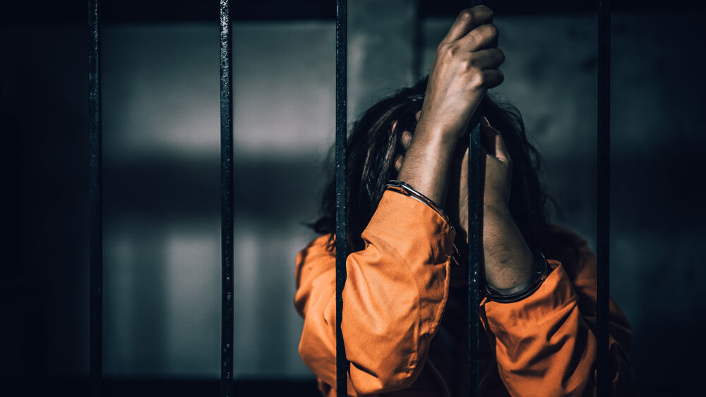 A woman holds onto the bars of her prison cell while wearing an orange jumpsuit.