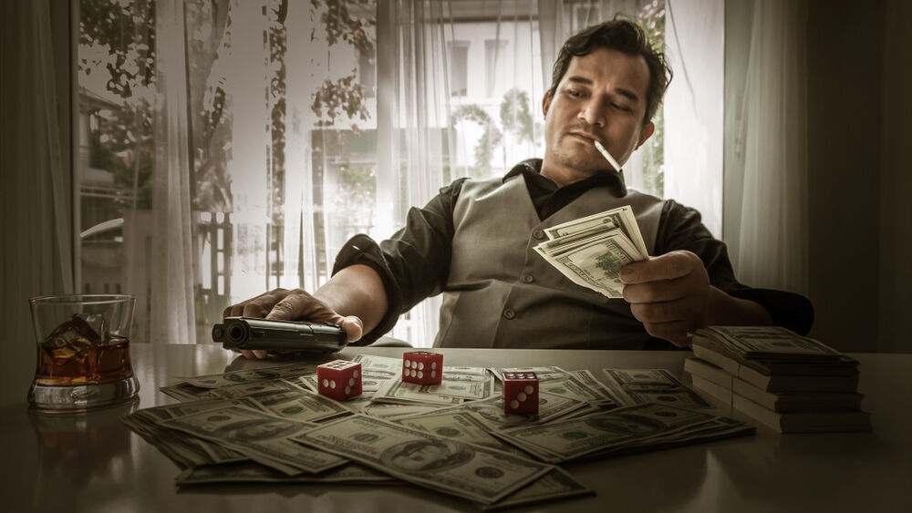 A mafia member counts his money while smoking and drinking and holding a gun.