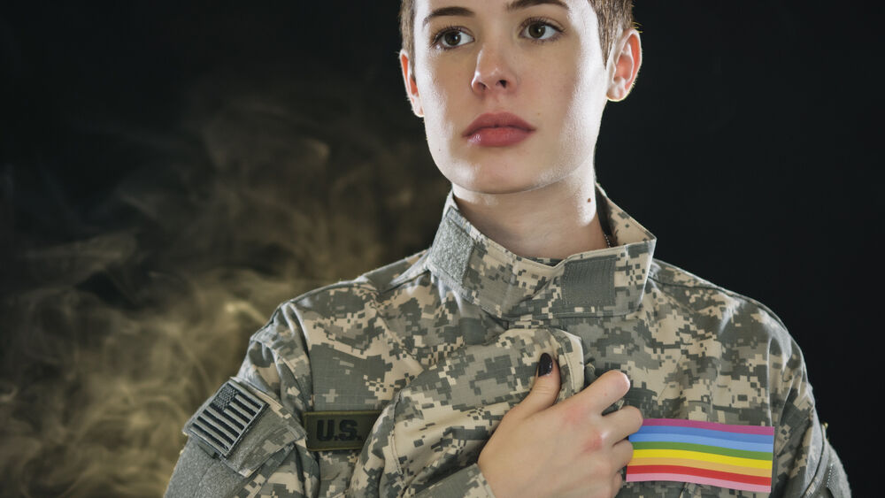 A queer soldier with a Pride flag on her uniform touches her heart.