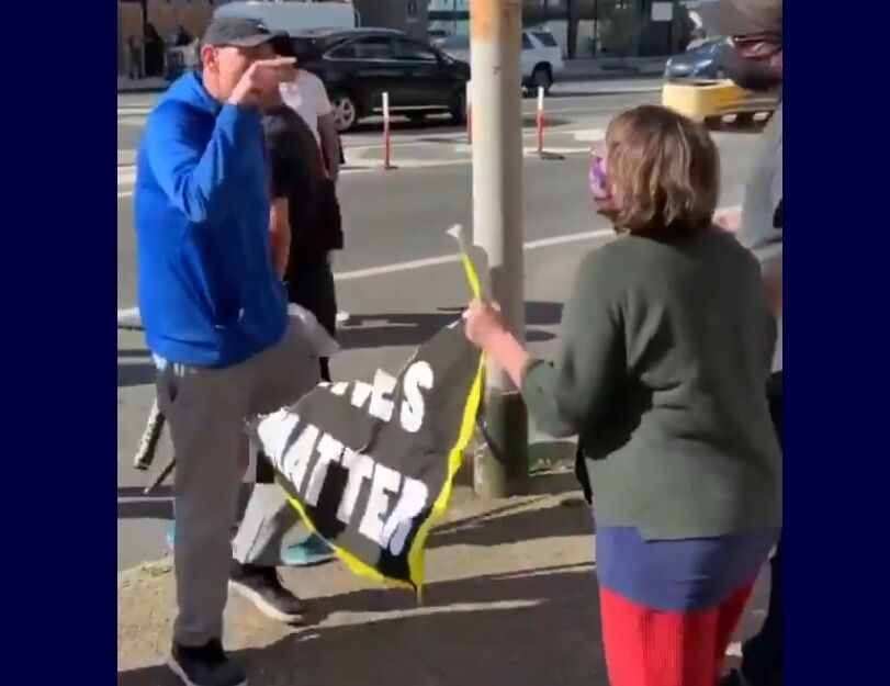 A man in a blue sweater pointing at a protestor with half a sign that formerly said "black lives matter"