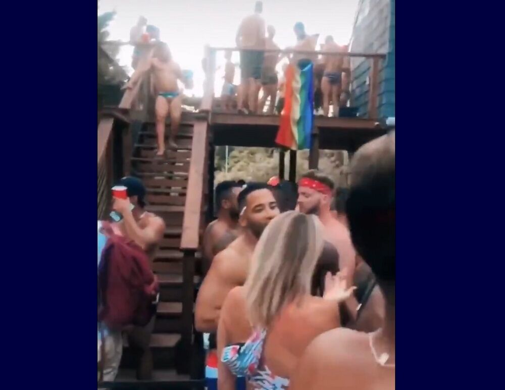 Shirtless partiers without masks and a rainbow flag in the background