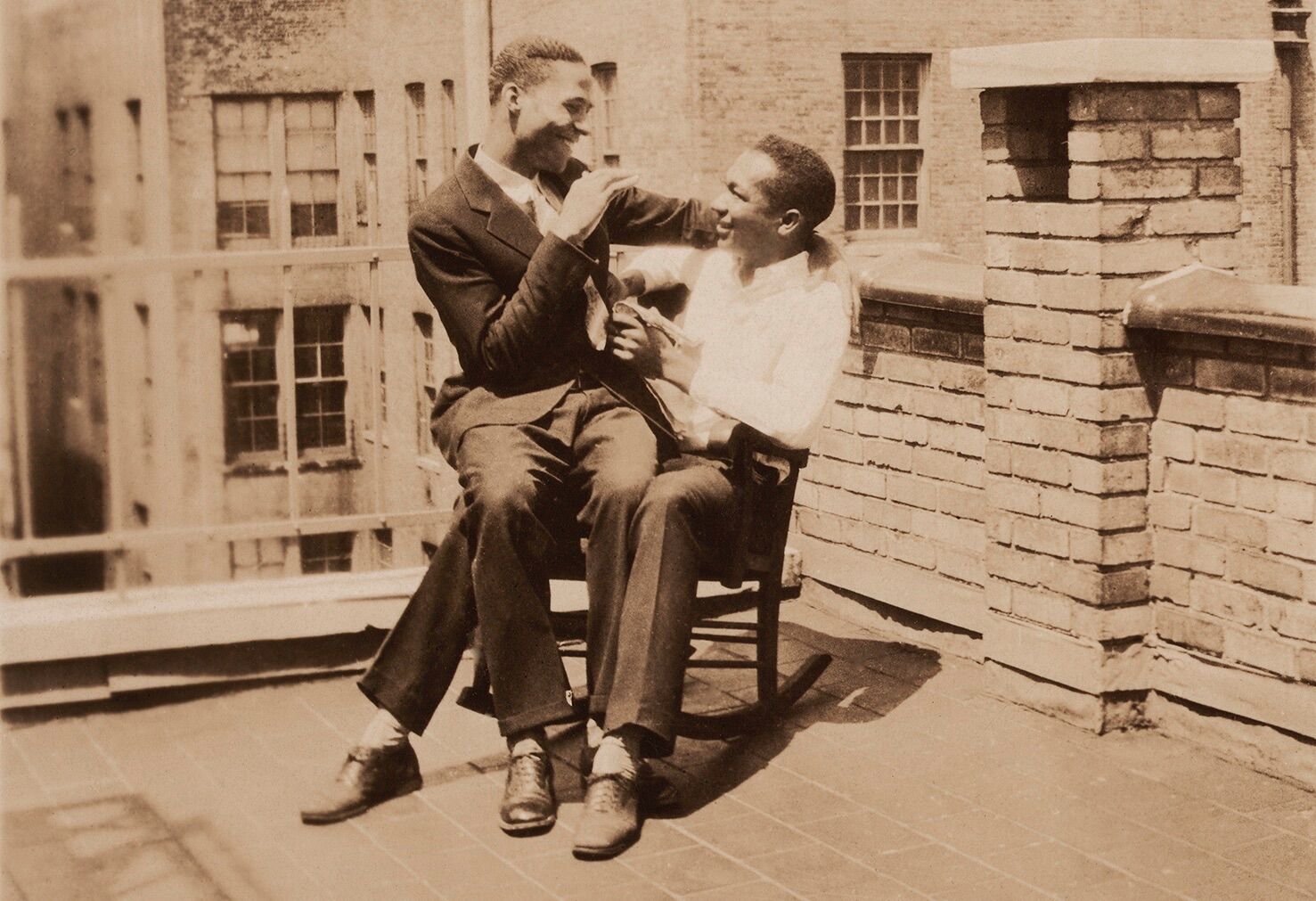 Two Black men pose in a rocking chair on a city rooftop