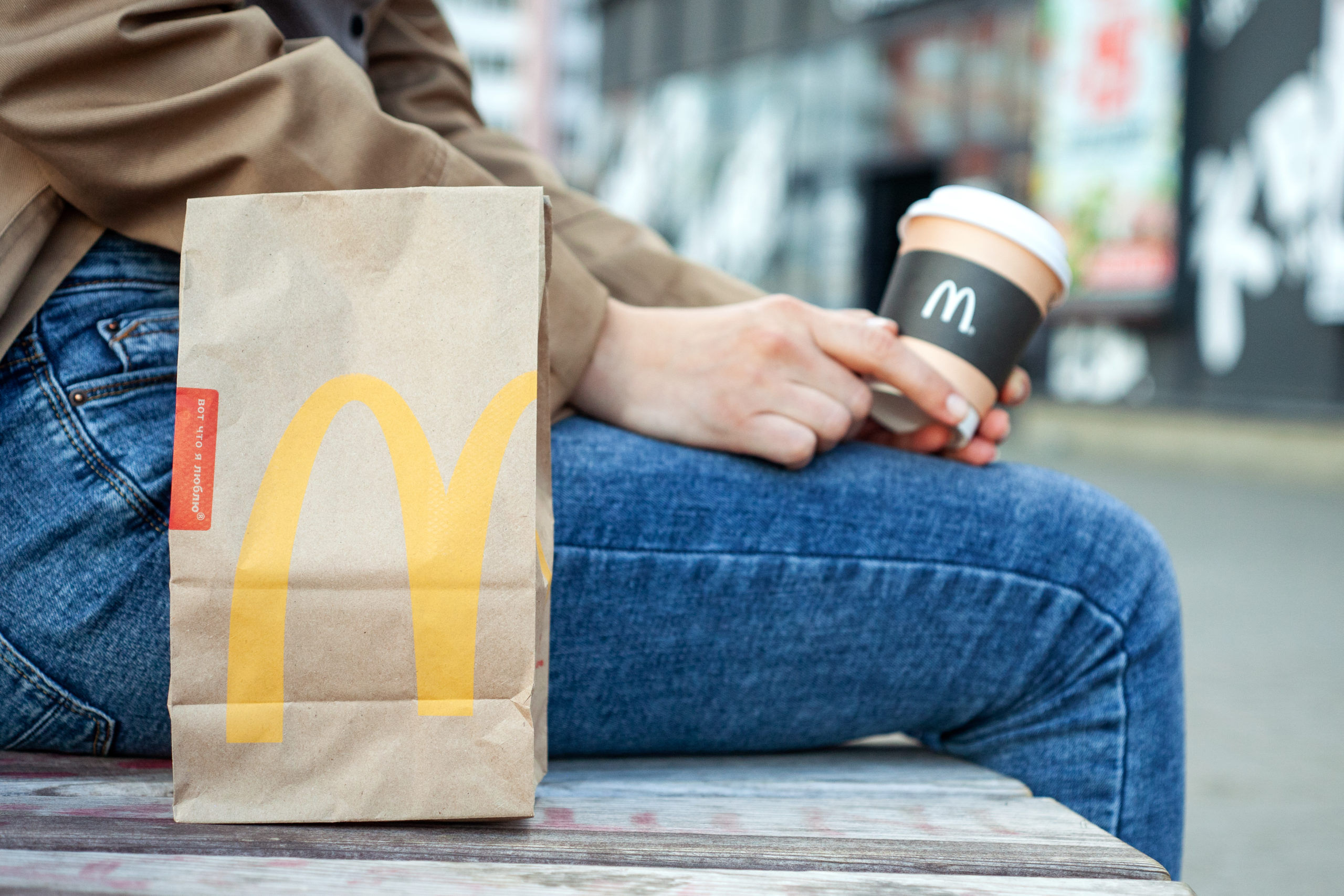 Minsk, Belarus, July 02, 2019: Takeout packing paper bag McDonald's on a bench next to a woman with paper cup of coffee in hand with McDonald's logo