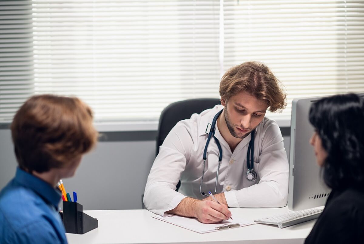 A doctor writing with a teen patient and another patient. Maybe the dishy doctor is writing a prescription for puberty blockers, as described in the article? It's a mystery... well, less a mystery and more like it's a stock photo