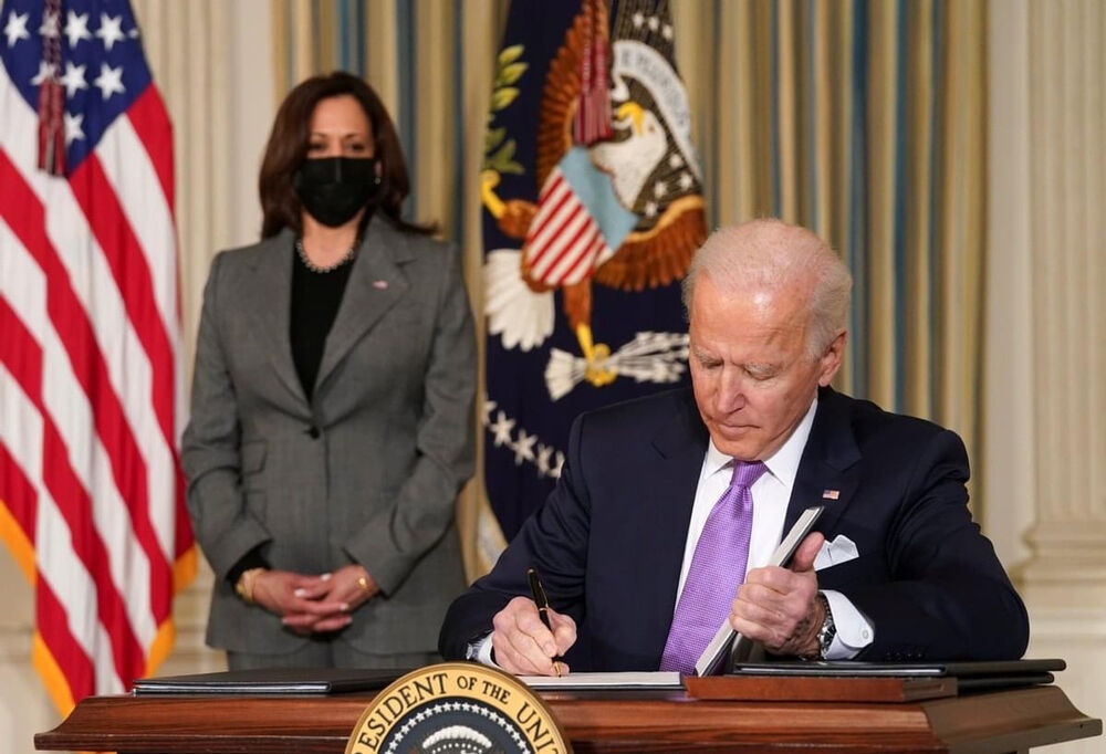 President Joe Biden signing papers in the White House as Vice President Kamala Harris looks on.