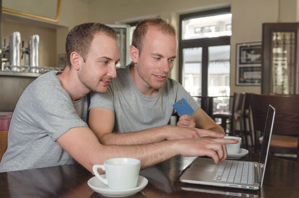 Gay couple using a credit card for something on a computer. They also have big mugs and are actually using saucers.