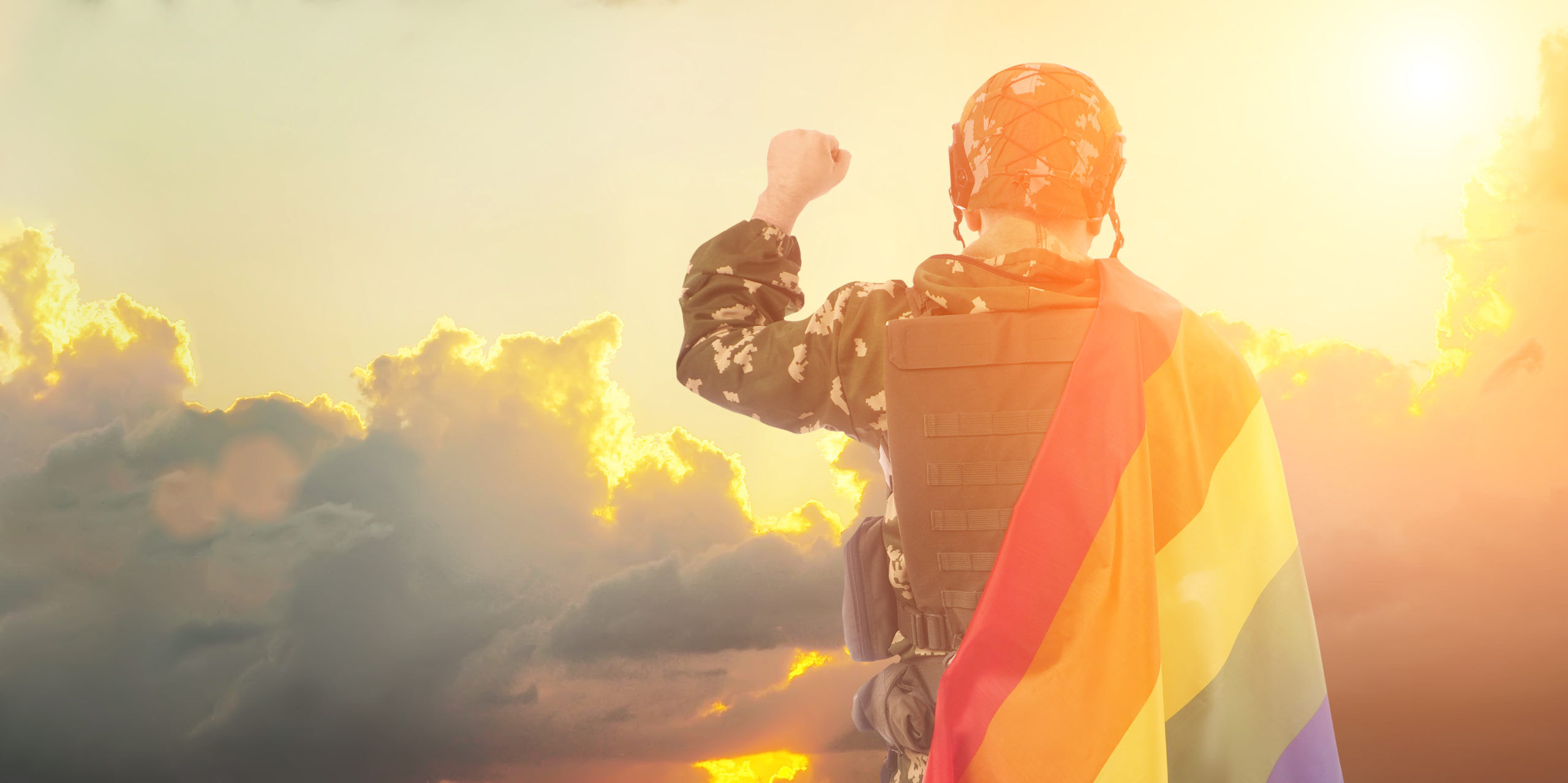 Member of military holding Pride flag under sunset