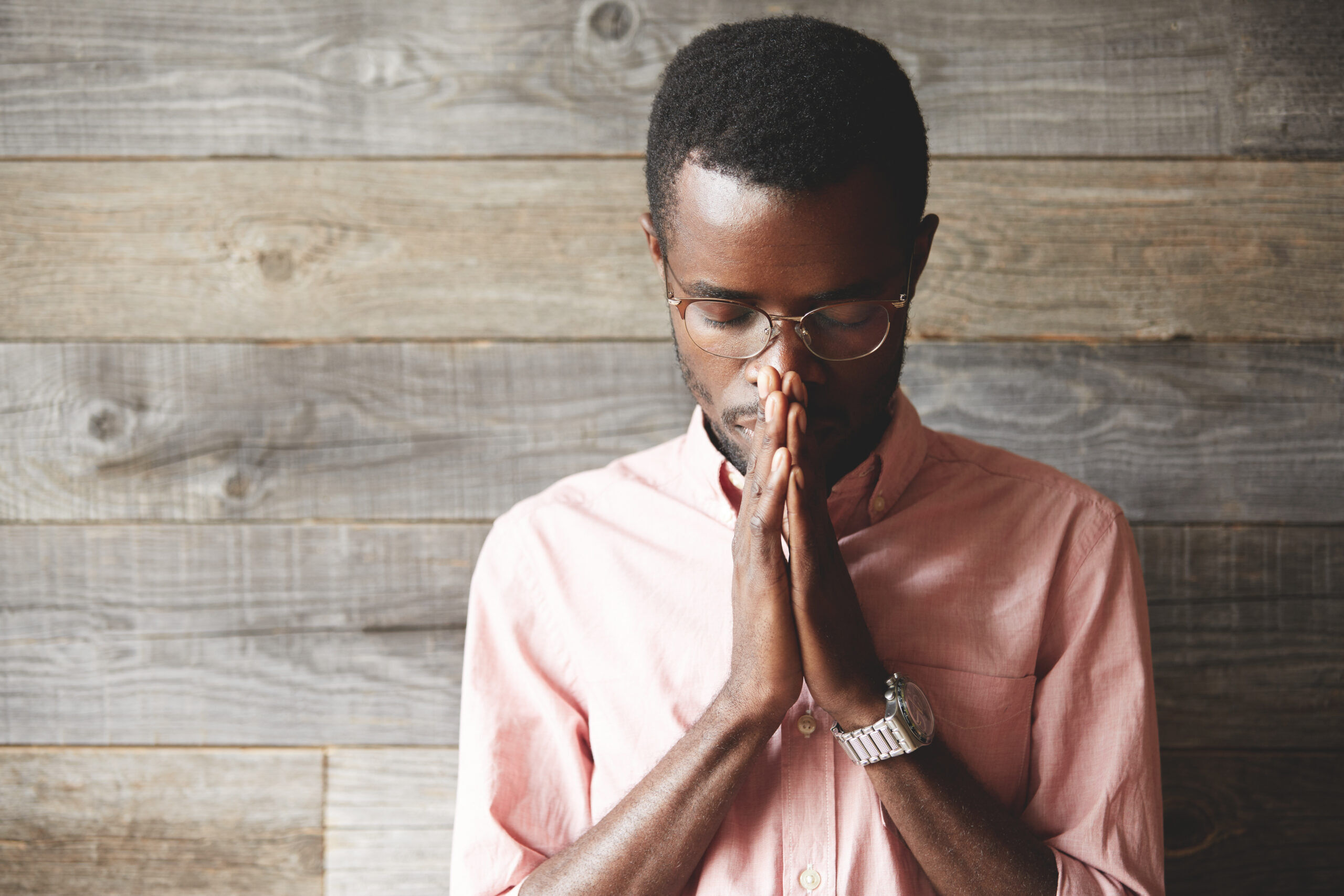 Young meditating and praying African American man wearing pink shirt and glasses, holding hands in prayer against his lips
