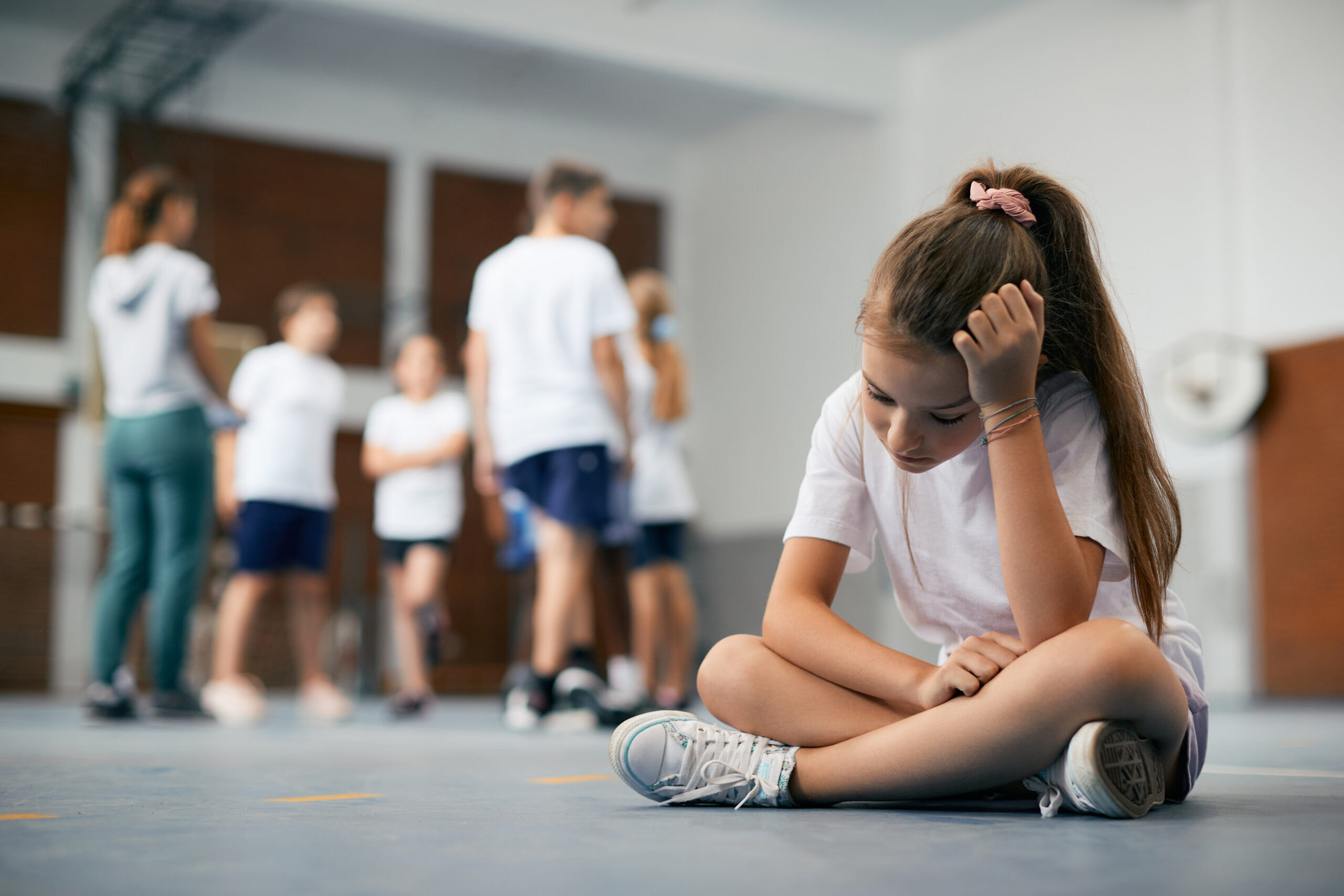 Elementary student sitting away from her classmates and teacher and feeling sad during physical education class.