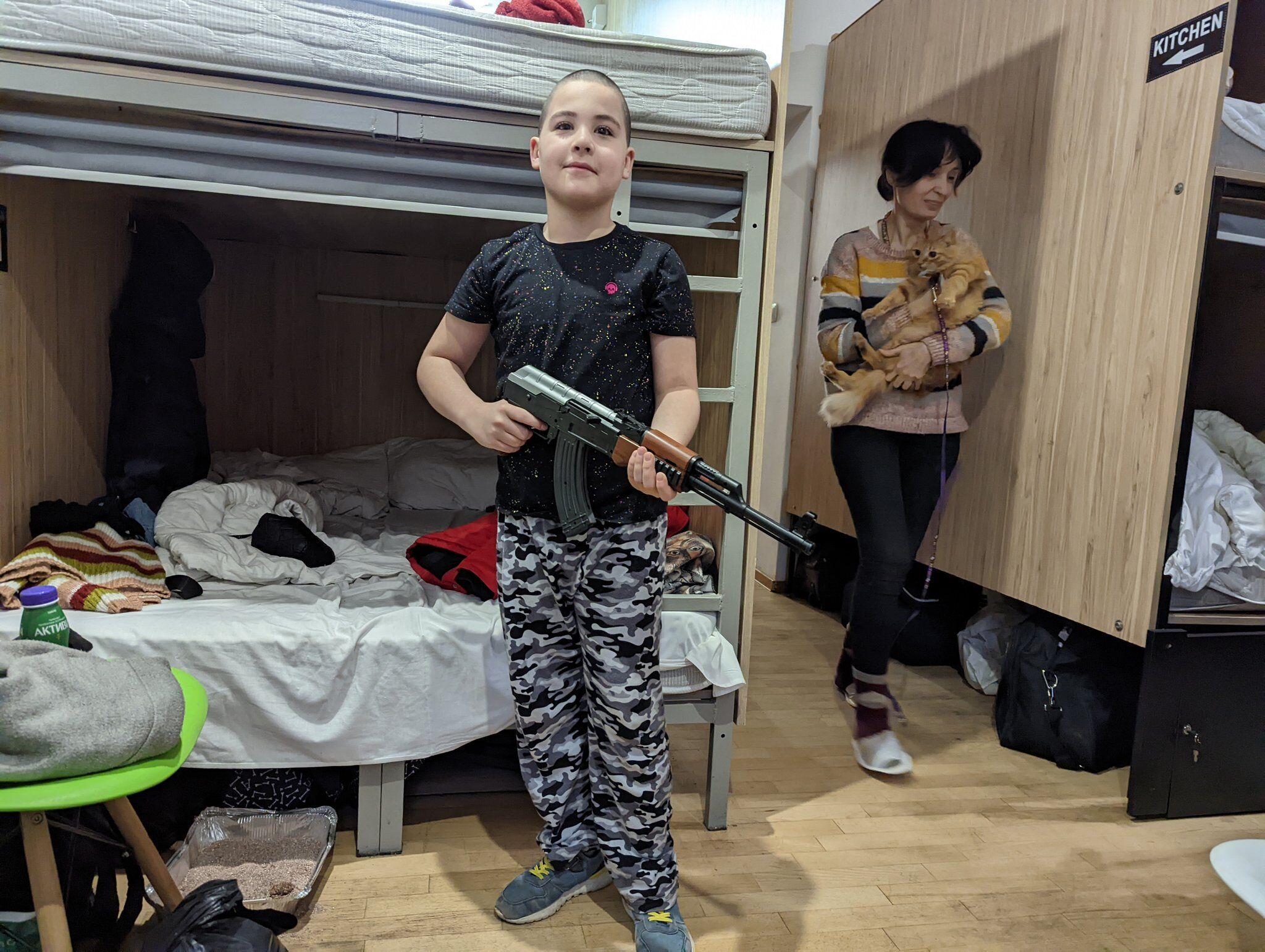 A boy poses with a gun as his mother walks by with the family cat. The two have been forced to flee their home as Russians invade Ukraine.