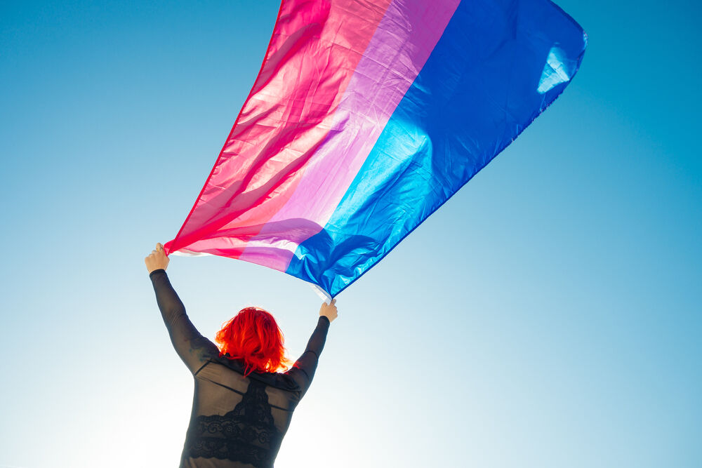 Woman with her back turned holding the bisexual flag flying above her head against a blue sky