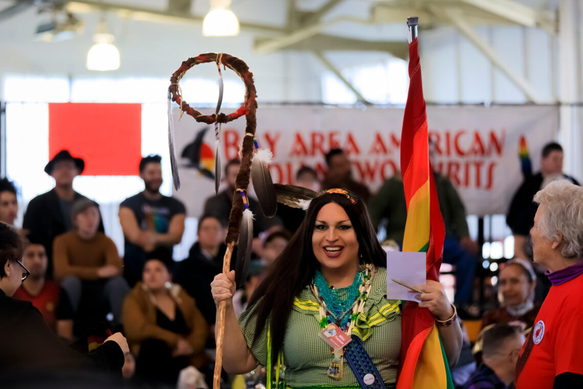 San Francisco, USA - February 08 2020: Native American Indian two spirit person holds a traditional Grand Entry eagle staff and rainbow flag at powwow