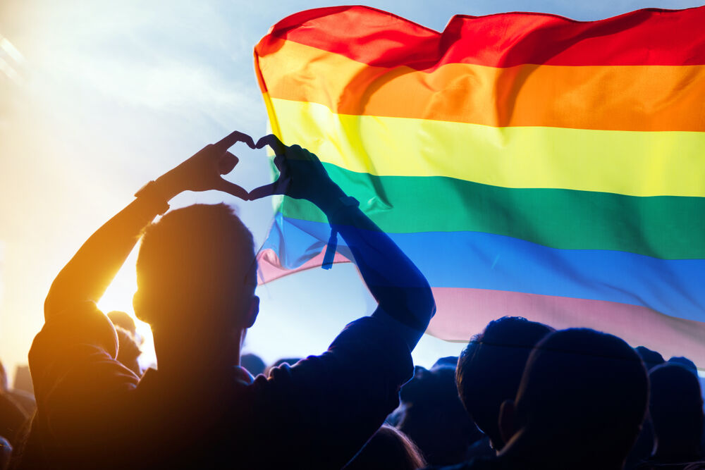 Silhoutte of a person making a heart with their fingers while staring at a Pride flag blowing in the wind against a blue sky