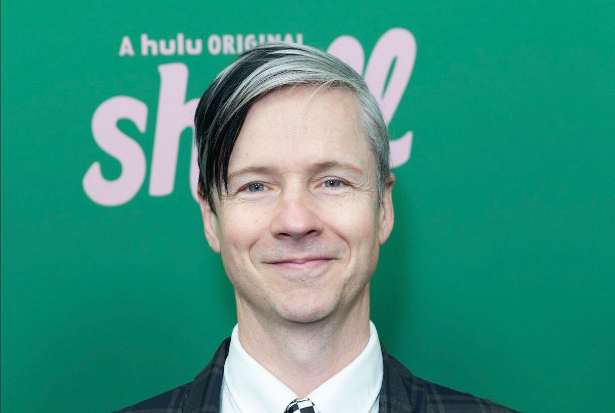 John Cameron Mitchell is a white man in his 60s who is wearing a collared shirt and tie and has grey streaks in his black hair. He is smiling against a green background.