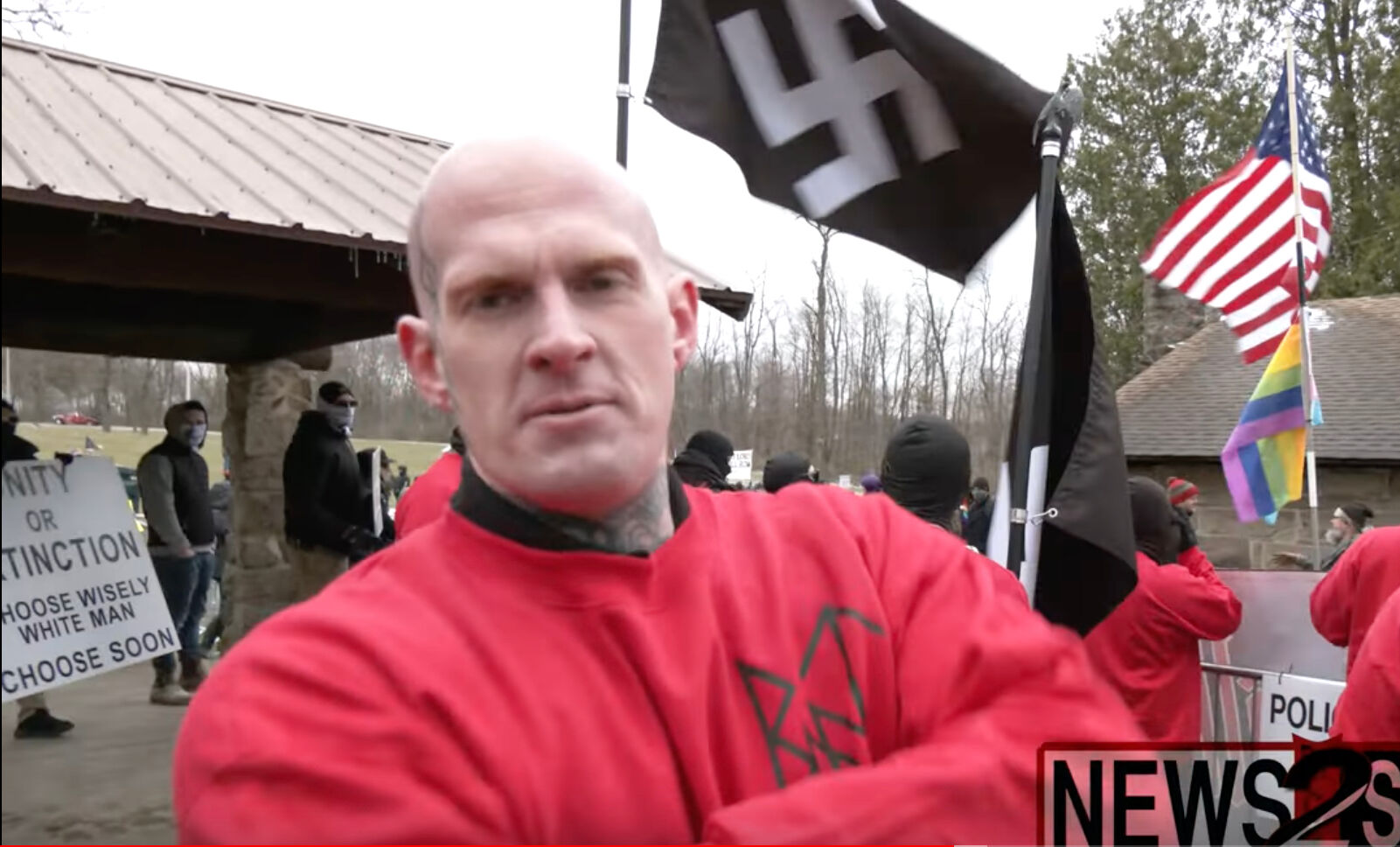 A bald white man in a red shirt stands near a park pavilion with a black Swastika flag flying behind him.