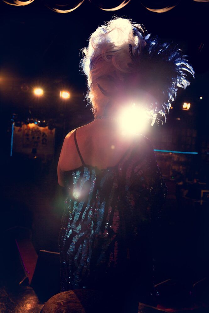 Darcelle XV spotlit from the back facing the crowd wearing big blue feathers in her hair and a blue sparkly dress