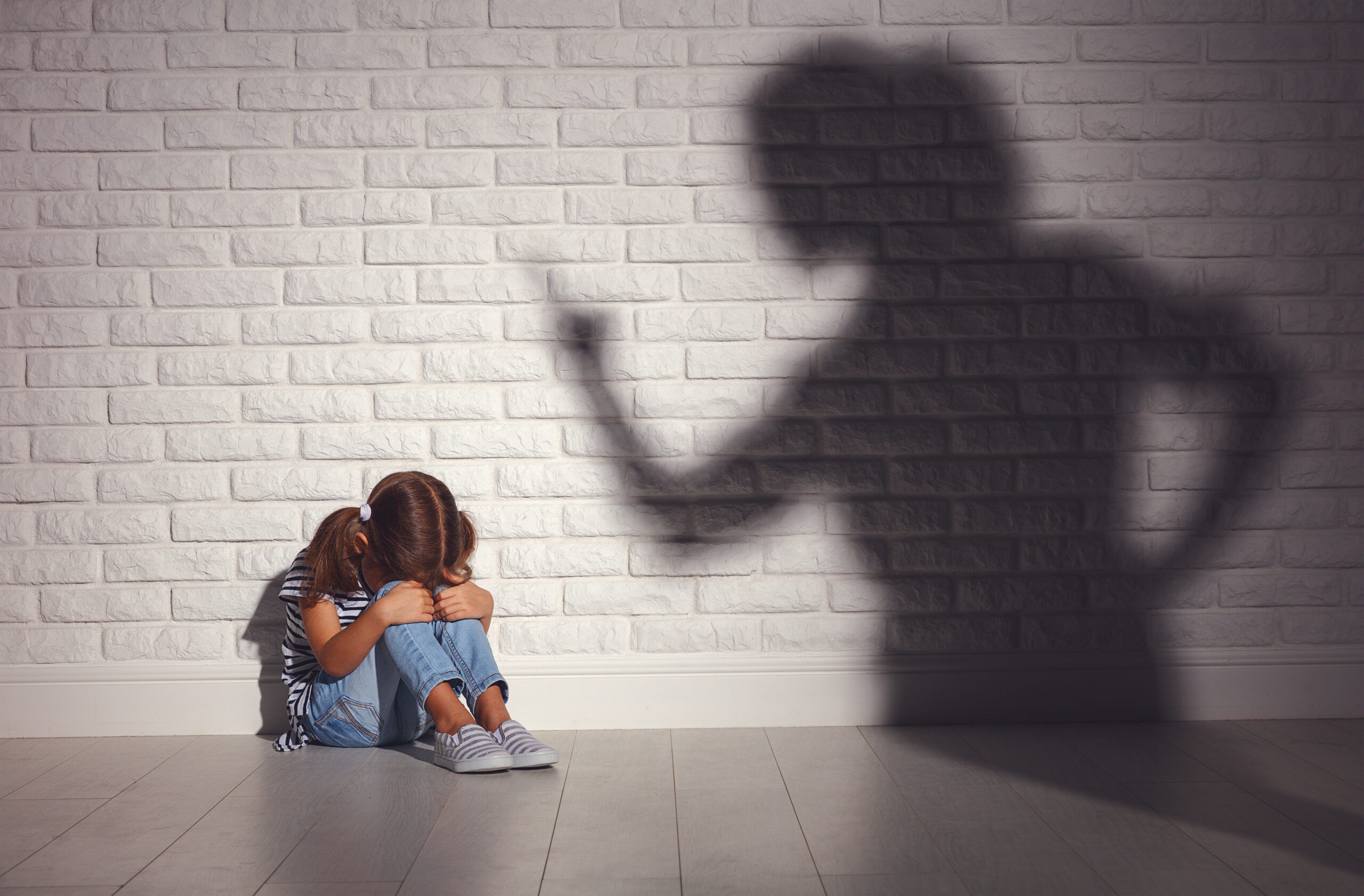 Shadow of an angry parent shouting at a sad kid who sits against a brick wall