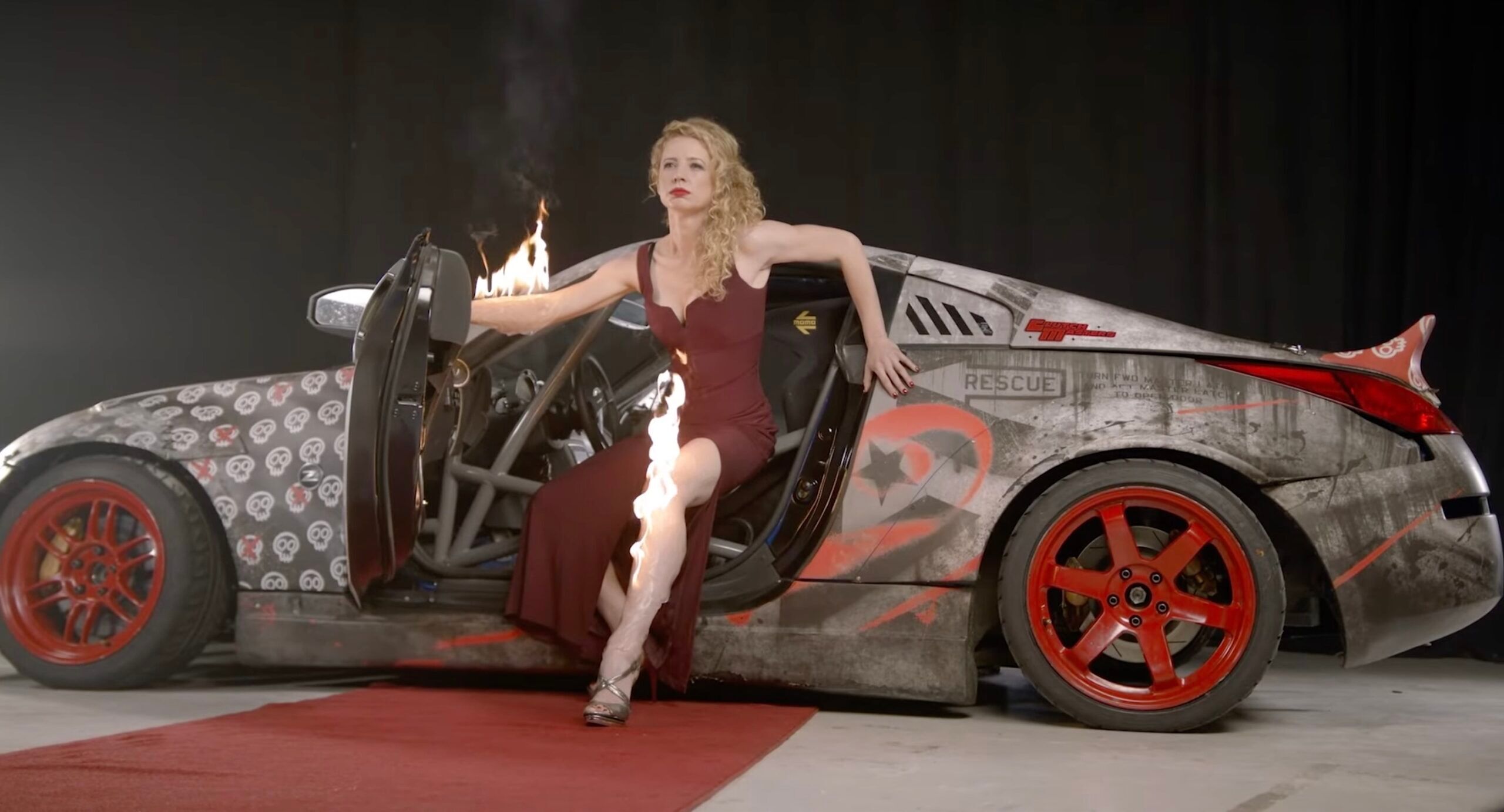 Queer stunt driver Zandara Kennedy poses with her arm and leg on fire in front of her drift car