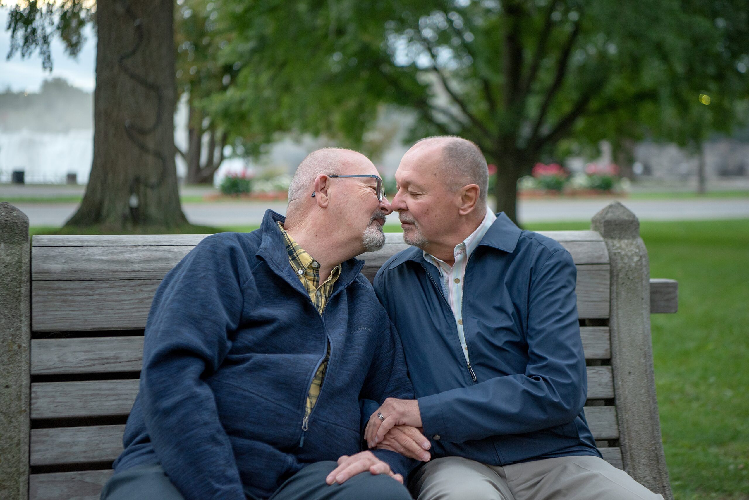 Elderly gay couple sitting on a park bench lean in to kiss each other. One has his arms linked through his partner's arms. Love and affection.
