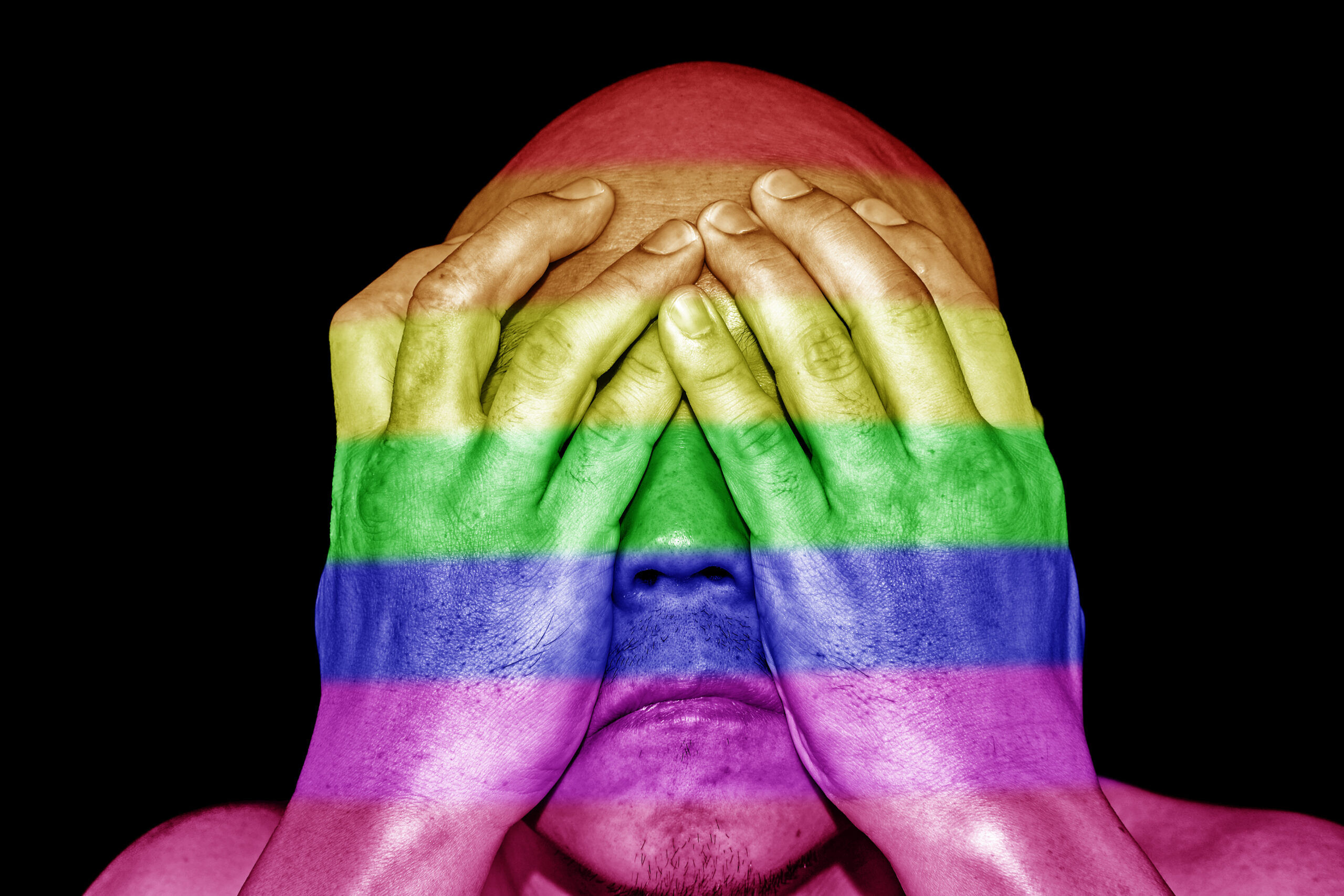 A man covering his eyes with his hands with imprint of rainbow colors on his skin