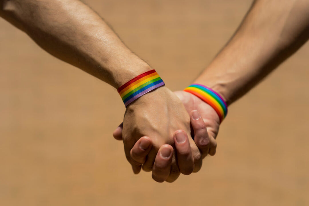 Close up of two hands holding each other, each wearing rainbow wristbands