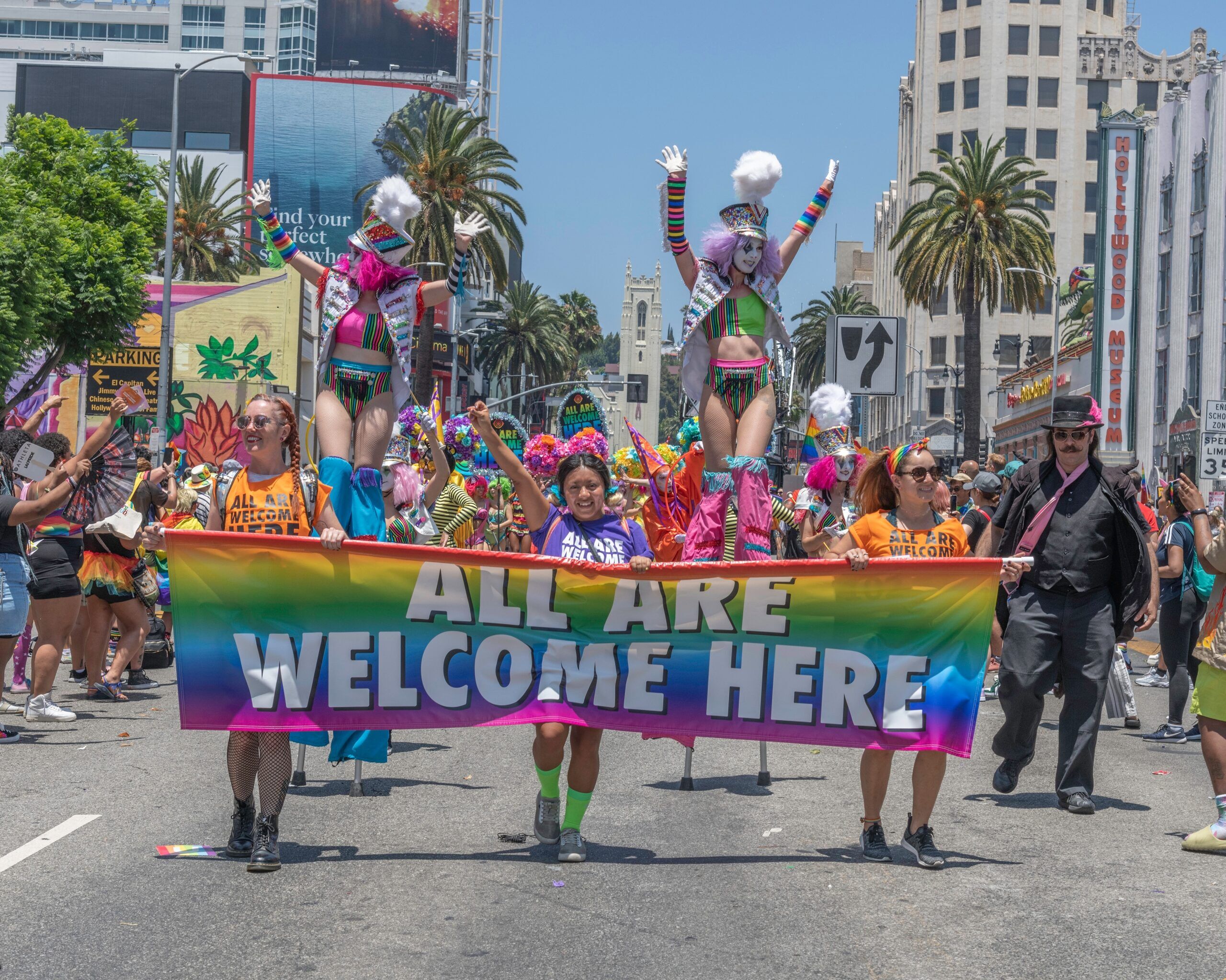 a colorful group of people march in the Los Angeles Pride Parade holding a sign that says "All are welcome here"