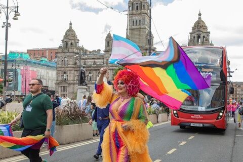 a drag queen wearing a colorful dress marches in the Glasgow Pride parade holding a Progress Pride flag in front of a double-decker bus