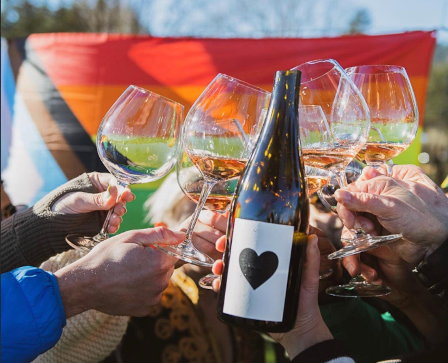 Queer Fine Fest, glasses clink with a bottle of wine with a heart label against a Pride flag