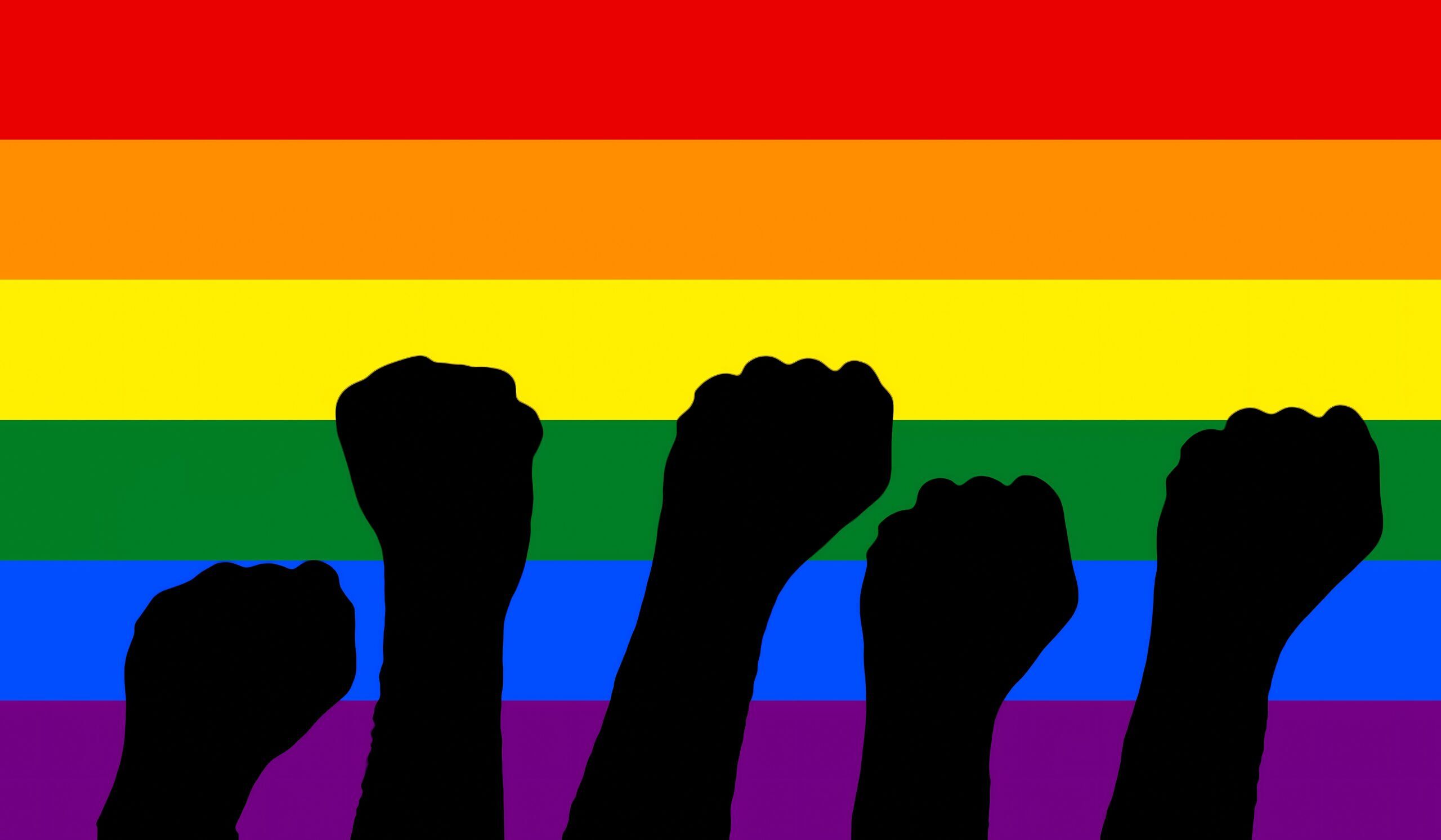 Fists raising (as dark silhouettes) over a rainbow LGBT flag. Symbols of protests and pride.