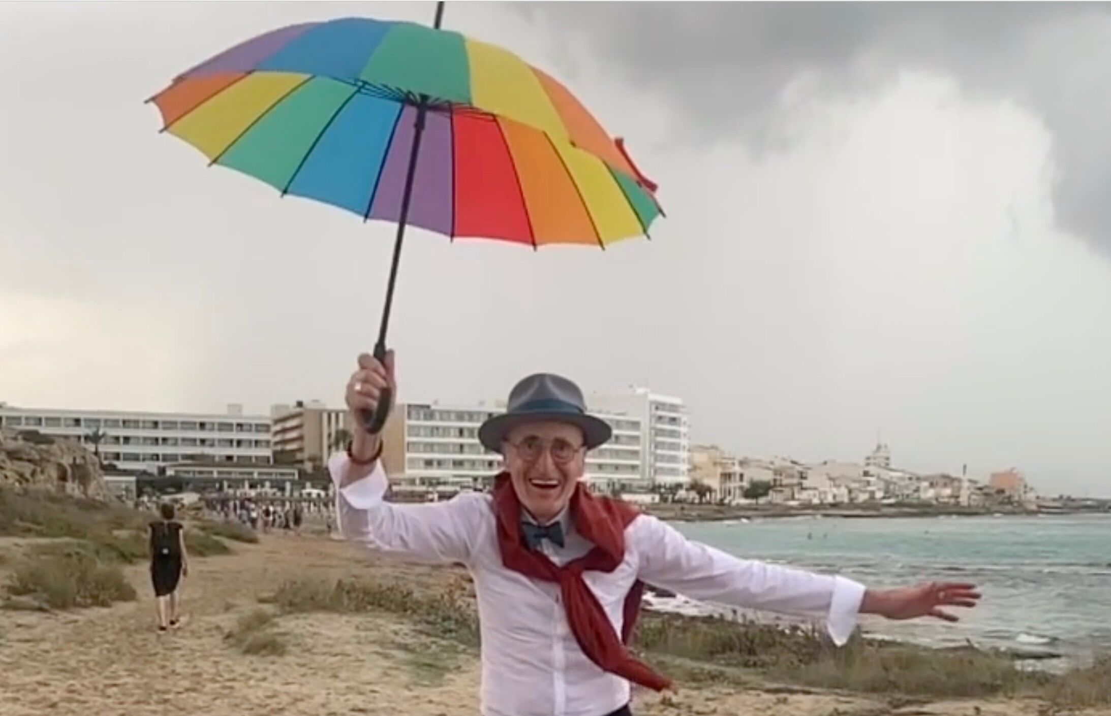 Photo of "hipster grandpa" Günther Krappenhofft dressed to the nines and jumping on the beach holding a rainbow umbrella