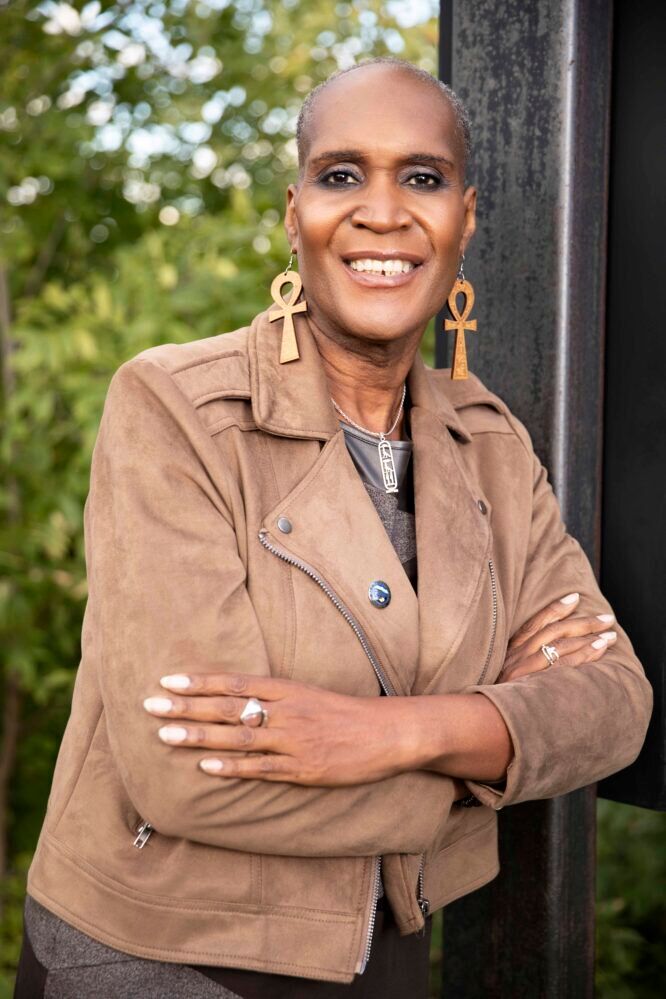 Andrea Jenkins smiling with her arms crossed wearing a jacket and large wooden earrings with greenery in the background