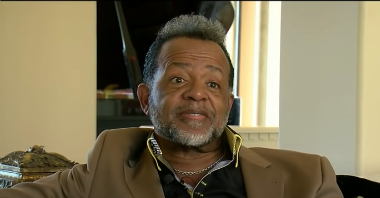 Carlton Pearson, a Black man with greyish curly hair, sits in a living room while wearing a dark open-collared shirt and a brown blazer