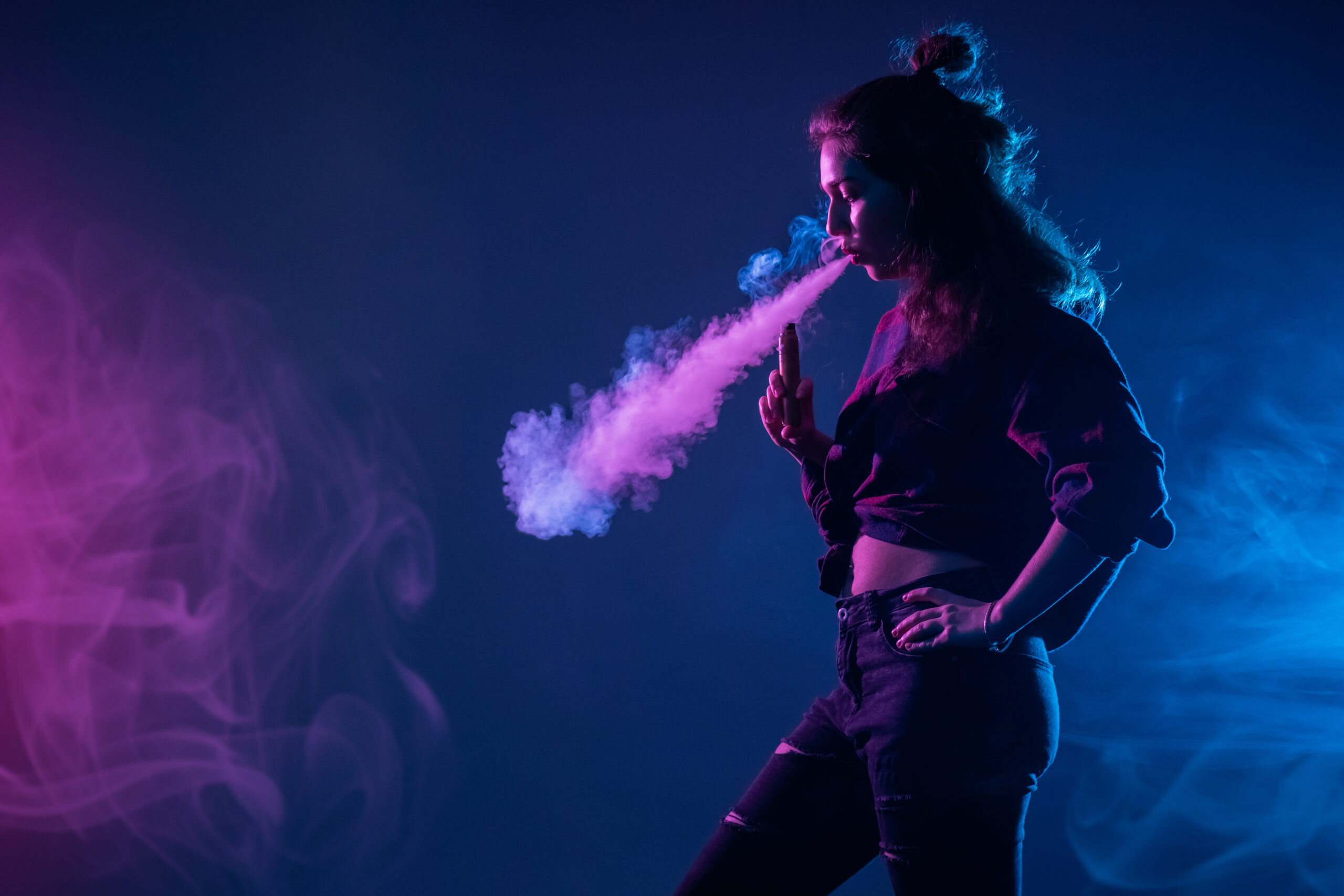 A young brunette woman in a black cut-off shirt and jeans blows out smoke while holding an electronic vape device in her hand. The light upon her is blue and purple.