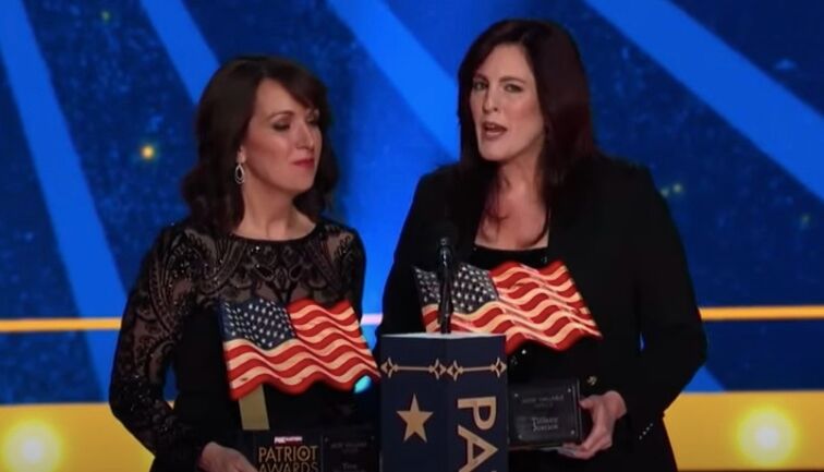 The Moms for Liberty co-founders hold two awards sculptures shaped like waving American flags while standing against a gold and blue background at Fox Nation's 2023 Patriot Awards